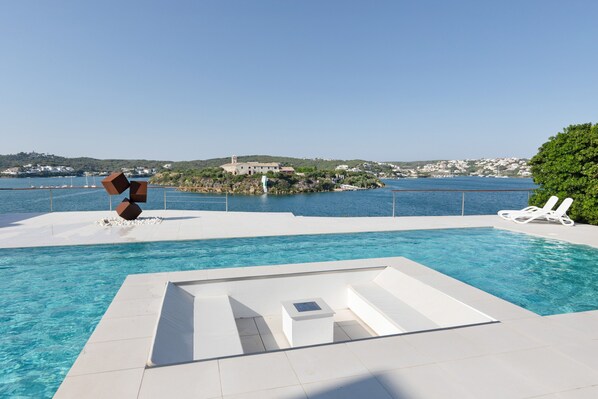 Una piscina con diseño geométrico, sillas de descanso blancas y vistas a un pueblo costero.