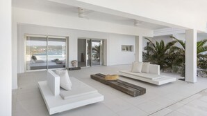 Un salon moderne avec des canapés blancs, un banc en bois et une vue sur la mer, visible à travers de grandes portes vitrées.