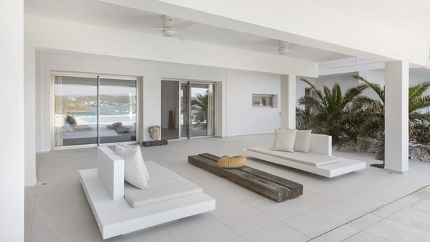 Un salon moderne avec des canapés blancs, un banc en bois et une vue sur la mer, visible à travers de grandes portes vitrées.