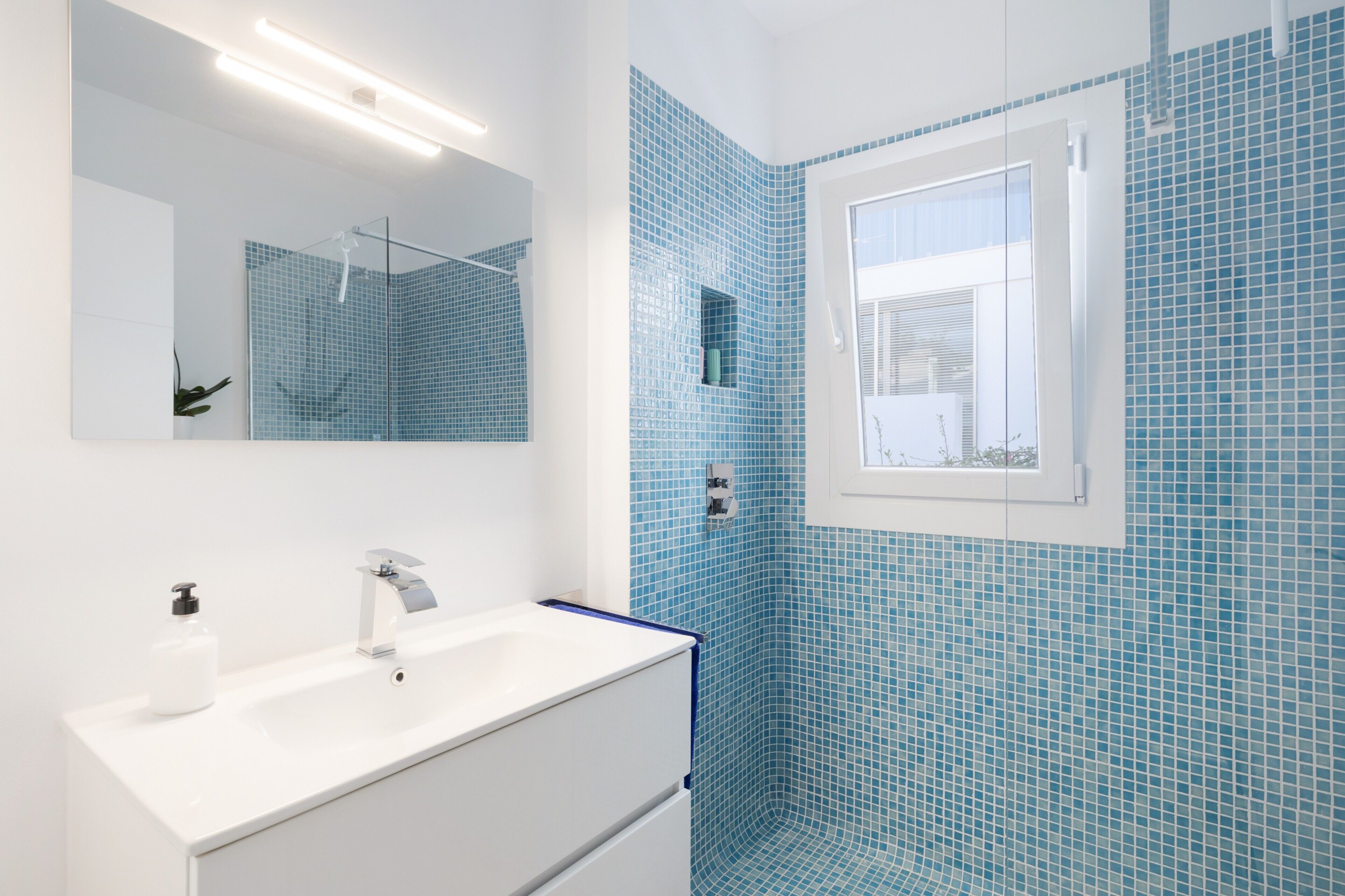 Un baño con lavamanos blanco, un espejo y una ducha con mosaicos azules.