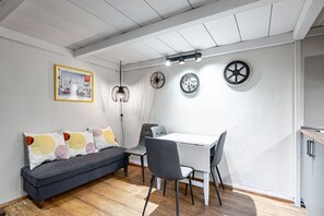 A modern kitchen with a white countertop, a dining table with chairs, a gray sofa with colorful cushions, and a ceiling with exposed beams and pendant lights.