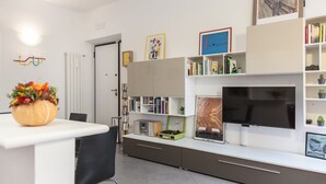 A modern, well-lit room with a white desk, a black chair, a bookshelf, a flat-screen TV, and a door.