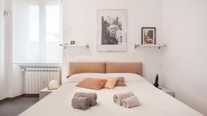 A hotel room with a bed, two pillows, a radiator, a window, and two wall-mounted shelves with framed pictures.