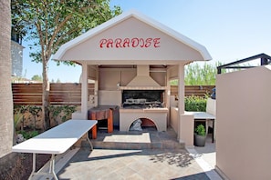 Ein Grillbereich im Freien mit einem 'PARADISE'-Schild, einem Kamin aus Stein und einem Tisch.
