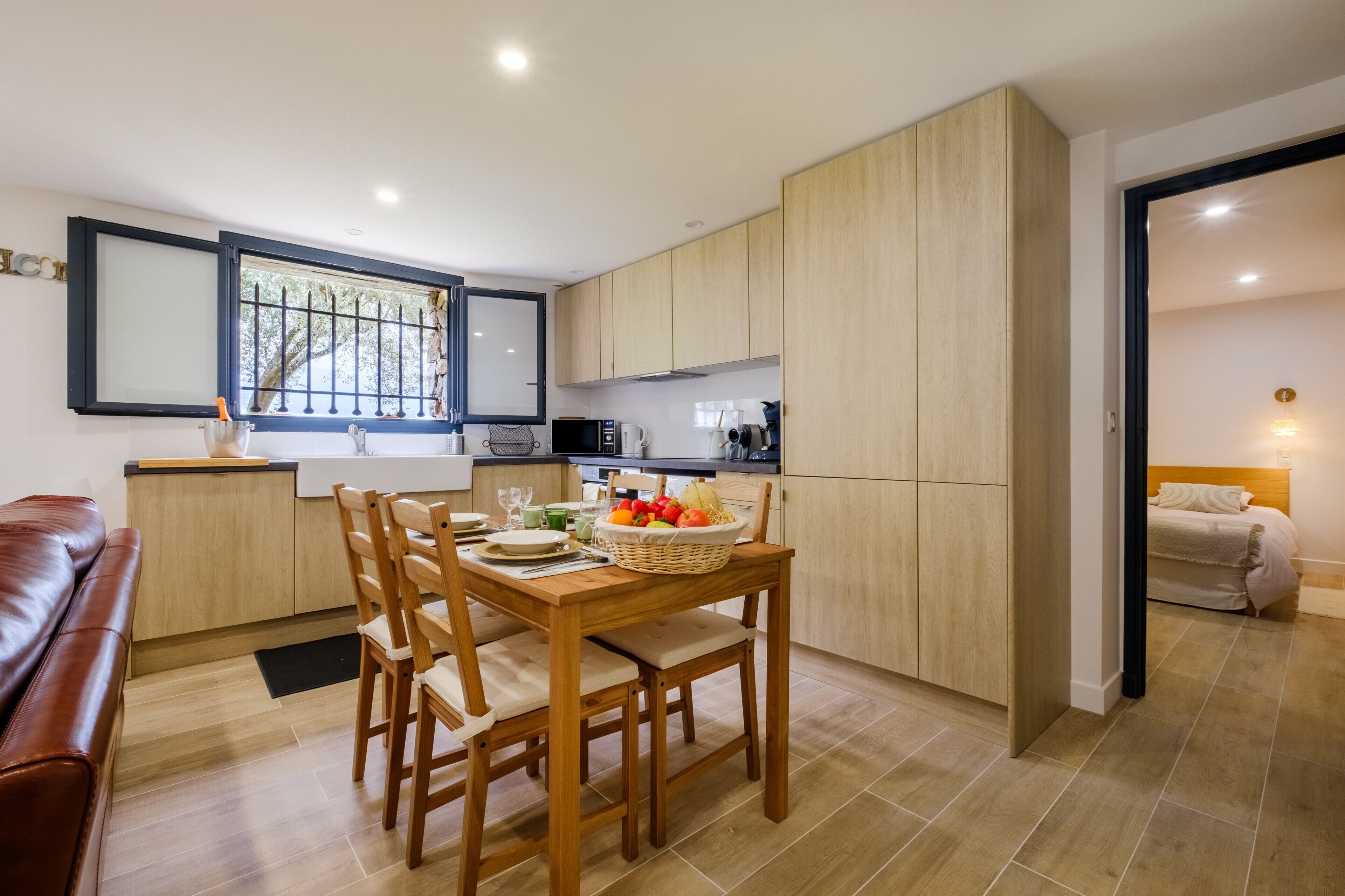 A modern kitchen with wooden cabinets, a dining table with chairs, a fruit basket, and a view through a window.