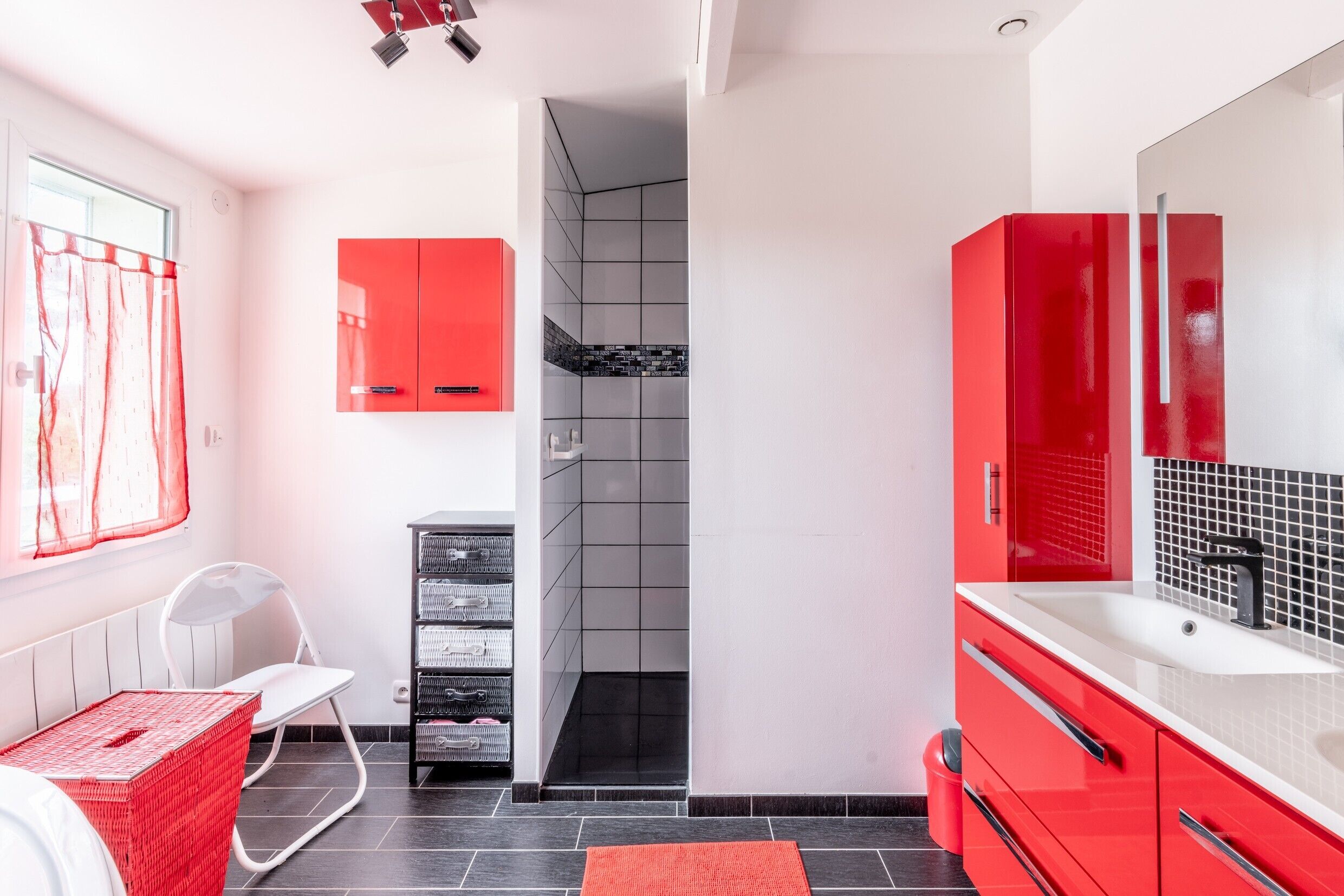Un baño moderno con un lavamanos rojo, armarios blancos y una zona de ducha con azulejos negros.