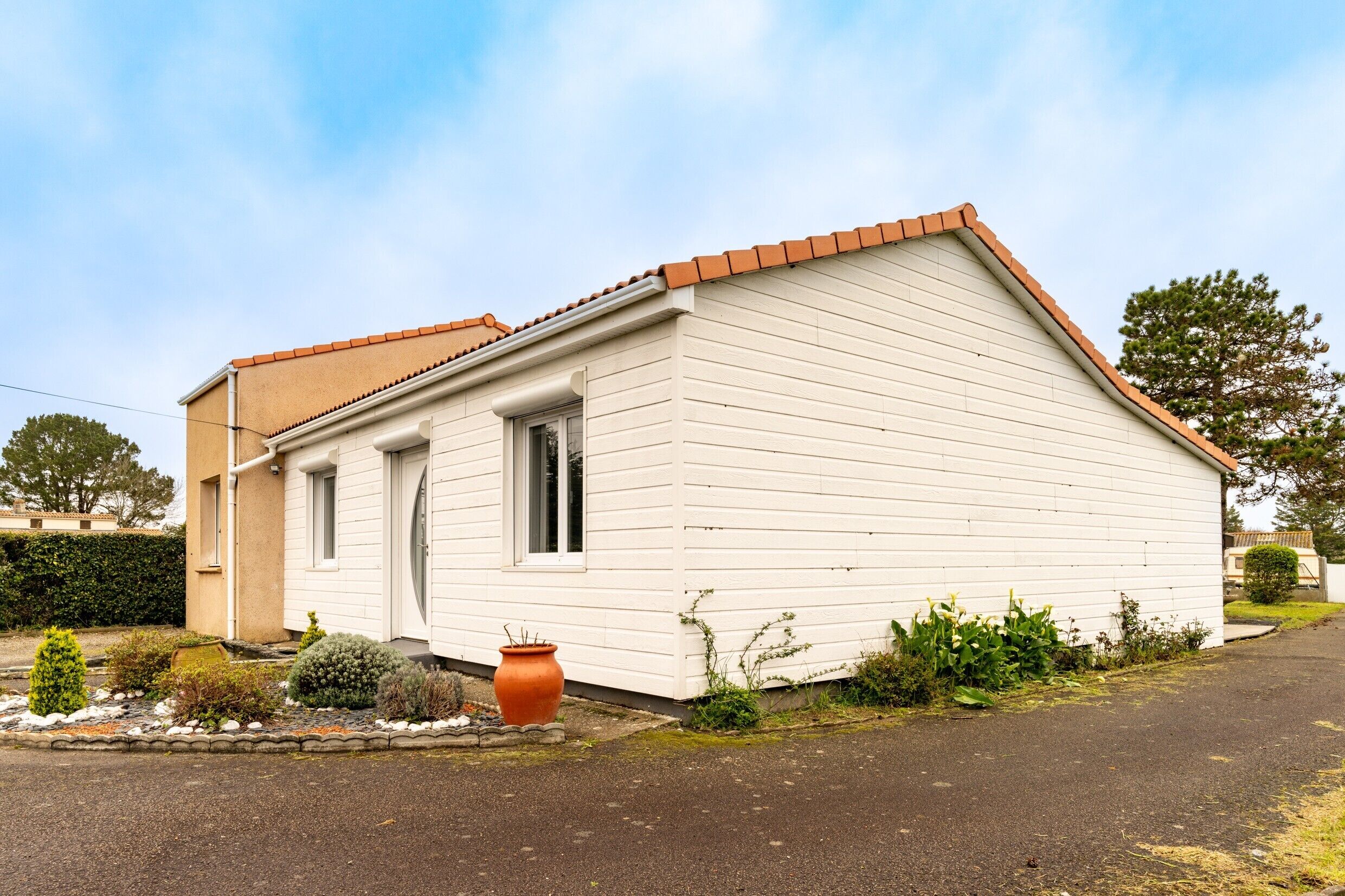 A single-story house with a sloped roof, a front door, and a small garden area with a large pot and various plants.