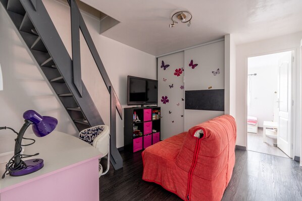 A modern living room with a staircase, a flat-screen TV, a pink desk with a purple lamp, a red sofa, and a bookshelf with pink storage boxes.