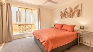Une chambre à coucher comprenant un lit, un ventilateur de plafond, une fenêtre avec des rideaux, une table de chevet avec une lampe et une œuvre d’art accrochée au mur.