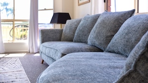 Un canapé d’angle gris dans une pièce bien éclairée, avec une grande fenêtre, un lampadaire noir et un tapis à motifs.