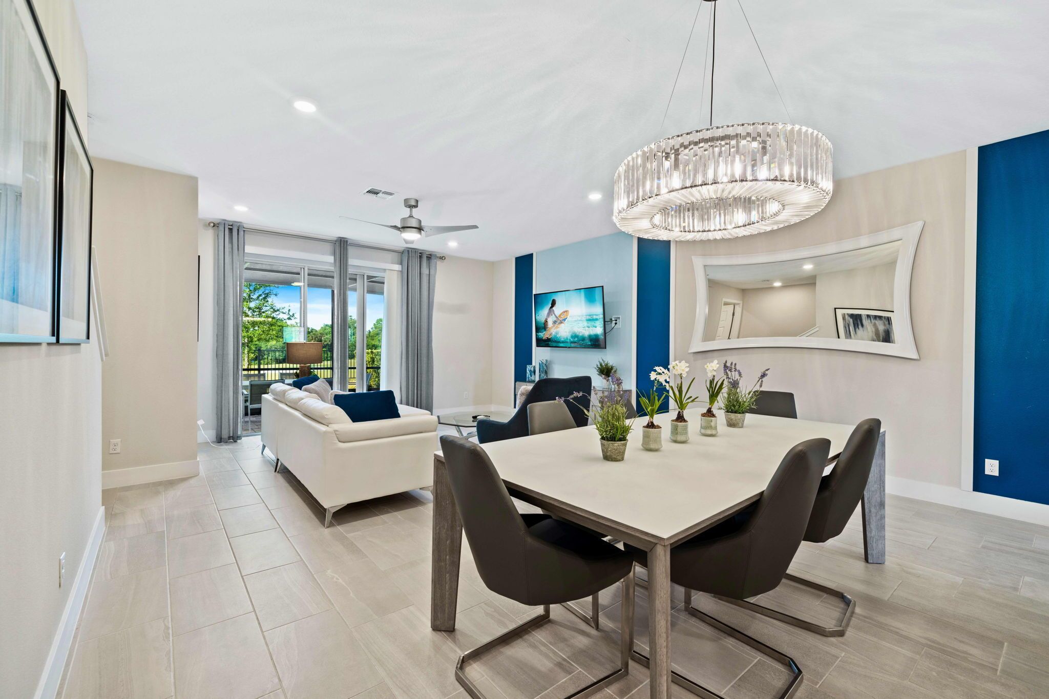Ein modernes Wohnzimmer mit einem weißen Sofa, einem Esstisch mit Stühlen, einer Kristallleuchtre und einem an der Wand befestigten Fernseher.