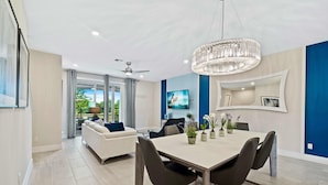 Uma sala de estar moderna com um sofá branco, uma mesa de jantar com cadeiras, um lustre e uma TV fixada na parede.