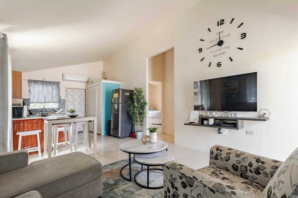 Un salon moderne avec une grande horloge murale, un téléviseur à écran plat et un coin repas, la cuisine étant visible au fond.