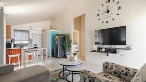 A modern living room with a large wall clock, a flat-screen TV, and a dining area with a kitchen in the background. - Tranquil Montego Bay City Getaway with Patio: The Perfect Stay! (Montego Bay)