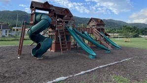 A wooden playground with slides and climbing structures, set against a backdrop of mountains and a clear sky. - Tranquil Montego Bay City Getaway with Patio: The Perfect Stay! (Montego Bay)