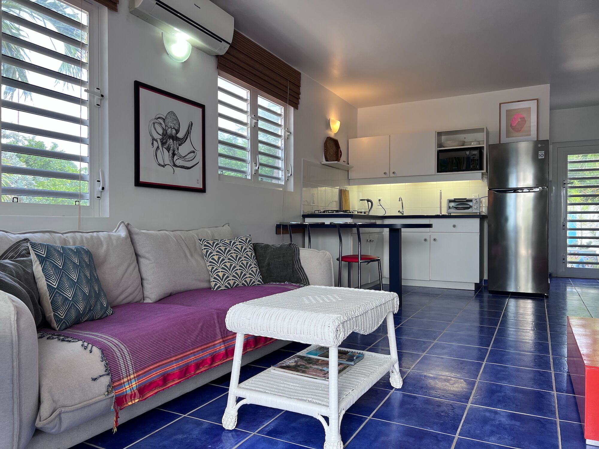 A modern living room with a sofa, a small white table, and a kitchen area with a refrigerator and cabinets.