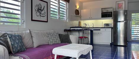 A modern living room with a sofa, a small white table, and a kitchen area with a refrigerator and cabinets.