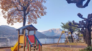 A playground with a slide, climbing steps, and swings, set against a backdrop of mountains and a body of water.