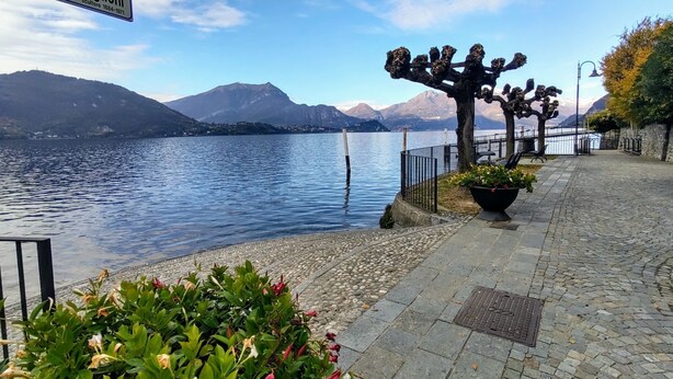A lakeside walkway with a sign reading "Lungolago Castiglioni," benches, potted plants, and a view of mountains.