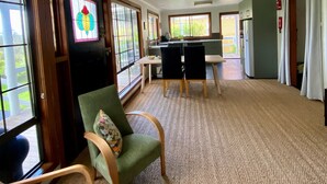 A room with a herringbone carpet, a green armchair with a floral cushion, a dining table with chairs, and a kitchen area with a refrigerator and stove.