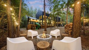 Una cabana moderna amb un espai foc i seients exteriors, envoltada d'arbres i llums penjants.