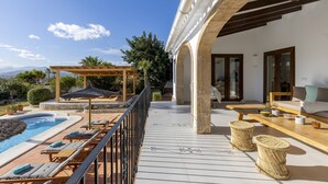Zona de piscina con sillones, una pérgola y una terraza de madera que conduce a un espacio de estar con sofá y mesa.