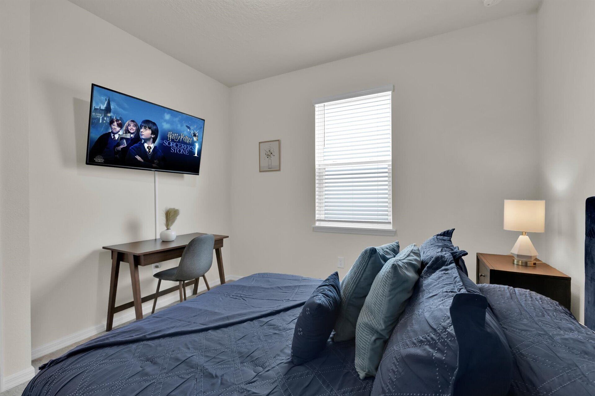 A bedroom with a bed, a desk, a chair, a TV, a window with blinds, and a bedside lamp.