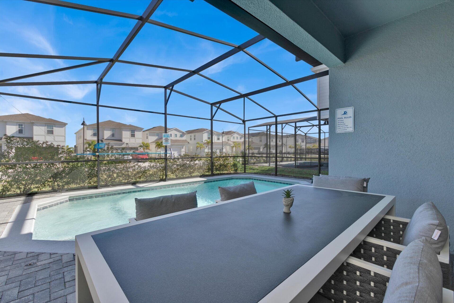 Un área de piscina exterior techada con mesa y sillas, rodeada por una estructura de metal y paneles de vidrio.