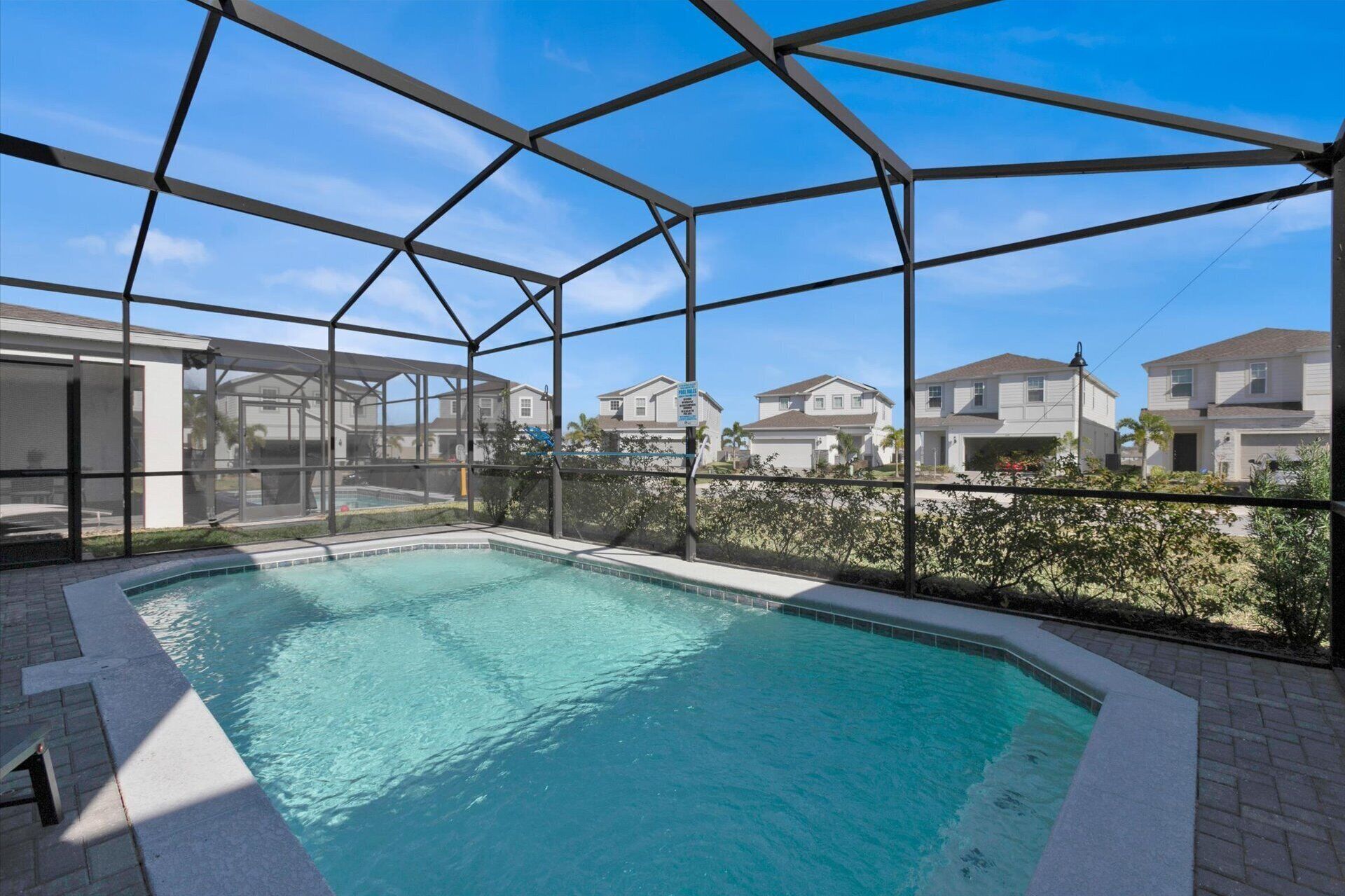 Une piscine dotée d’une structure de toit en métal, entourée d’une terrasse en briques et offrant une vue sur des maisons résidentielles.