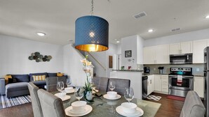 A modern kitchen with stainless steel appliances, a dining area with a glass table, and a blue hanging light fixture. - West Lucaya Village 4 Bedroom Townhome (Kissimmee)