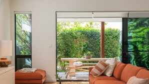 Una sala de estar moderna con sofá, reposapiés y mesa de centro. Hay una puerta corrediza de vidrio que da a un jardín con mesa y sillas de madera.