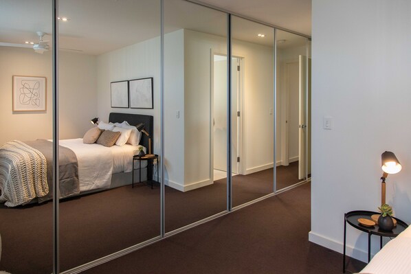 A modern bedroom with a large mirror, a bed with a headboard, bedside tables, and a lamp.