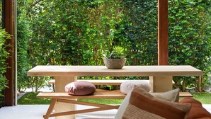 Una mesa de madera con una planta en maceta, un banco y cojines en una habitación con ventanas amplias que dan a un jardín exuberante.