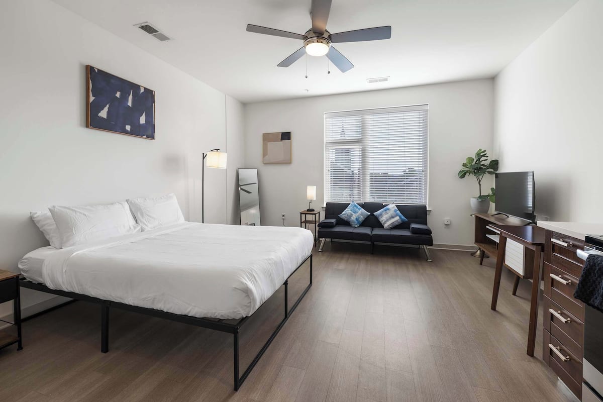 Une chambre à coucher comprenant un lit, un bureau, une télévision, un ventilateur de plafond, une fenêtre avec des stores, une plante et des œuvres d’art accrochées au mur.