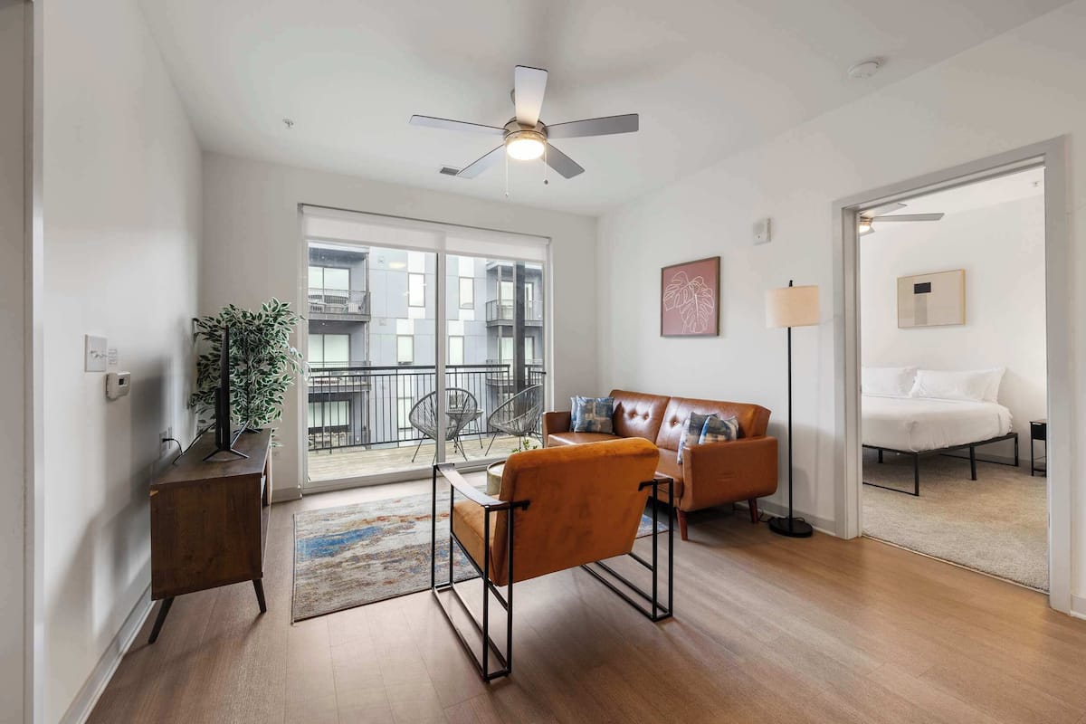 Un salon moderne avec un canapé, un fauteuil, une petite table et un ventilateur de plafond. On aperçoit un balcon à travers la porte vitrée.