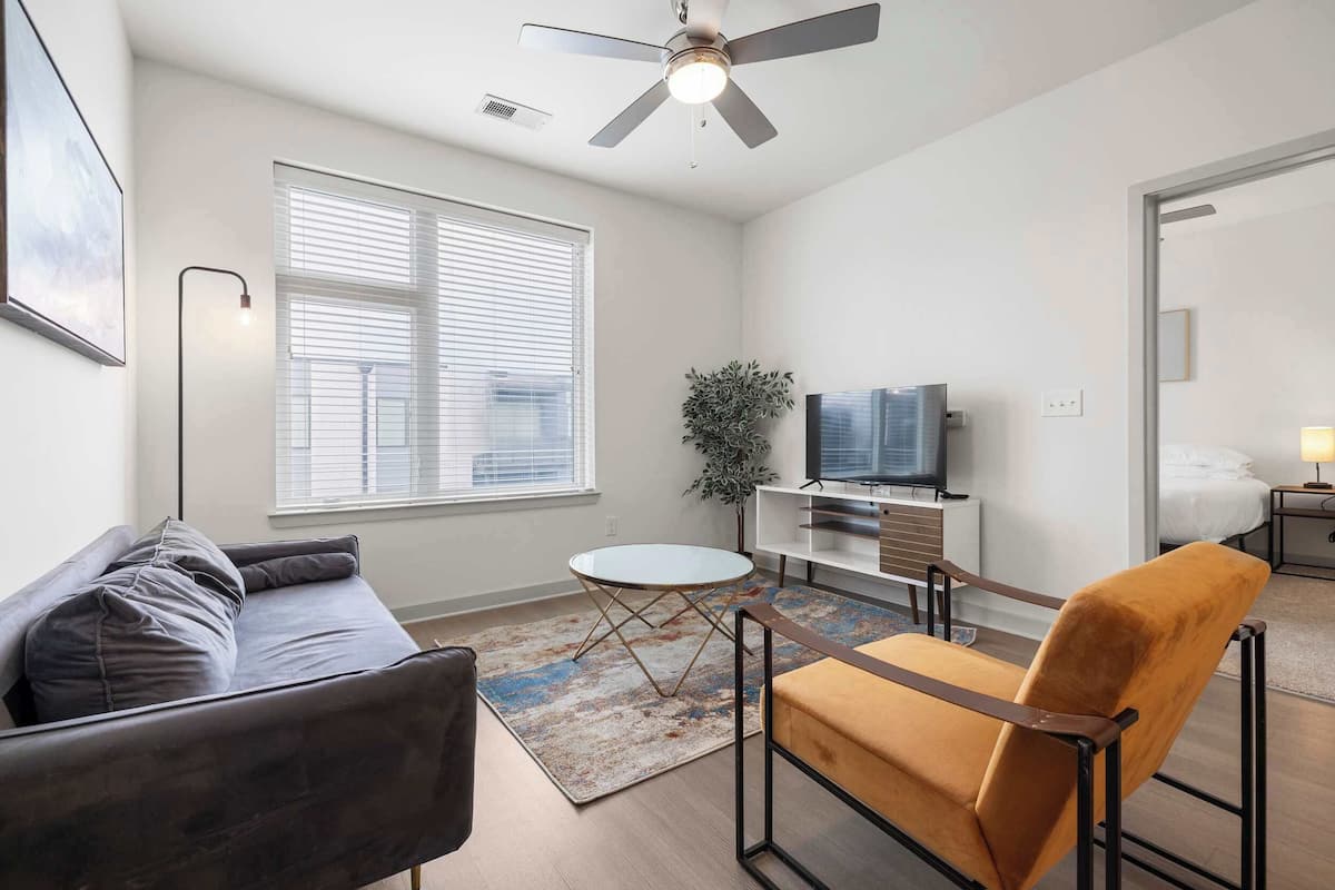 Un salon moderne avec un canapé, un fauteuil, un téléviseur et un ventilateur de plafond.