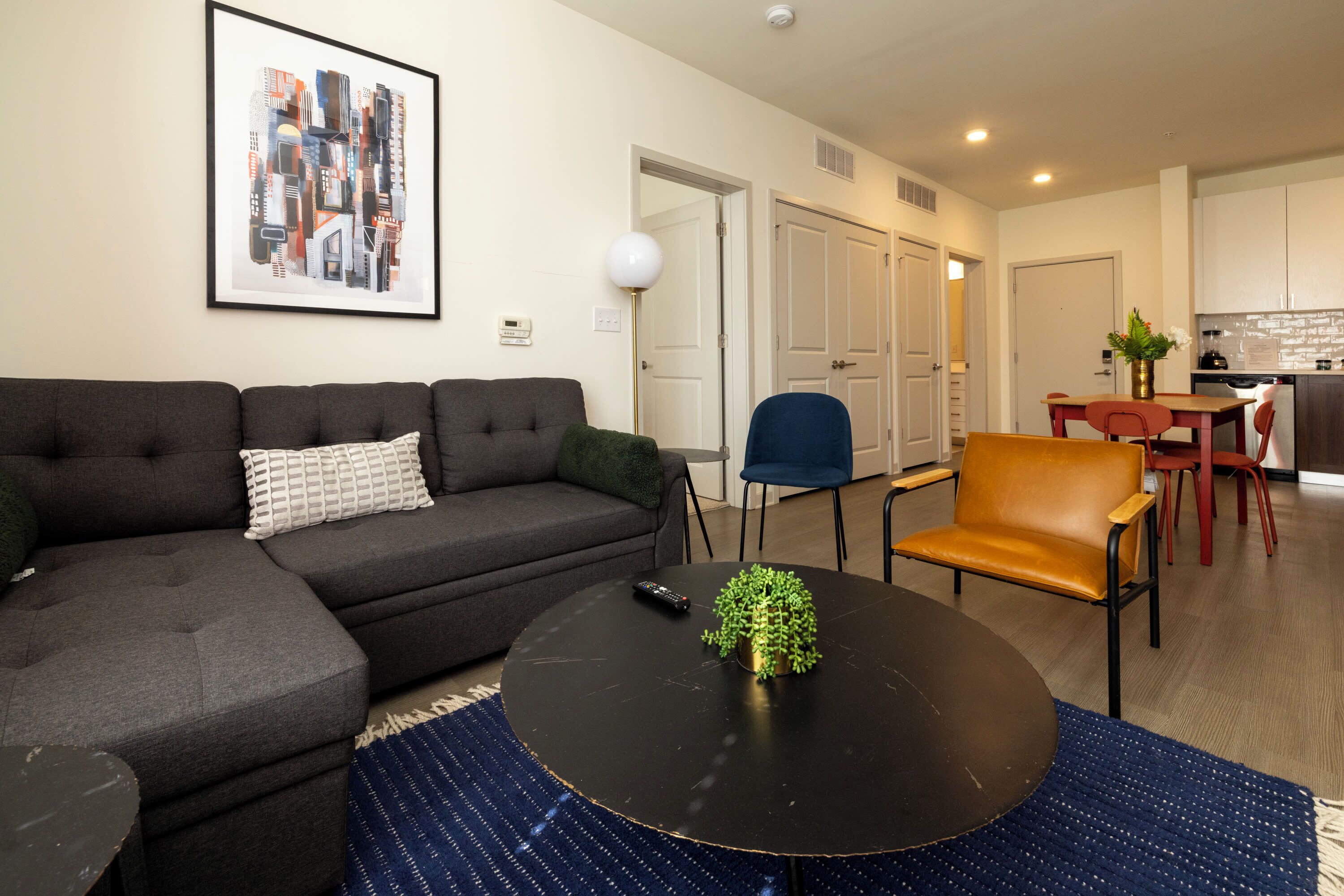 A living room with a sofa, a coffee table with a plant, a dining area with chairs, and a kitchen in the background.