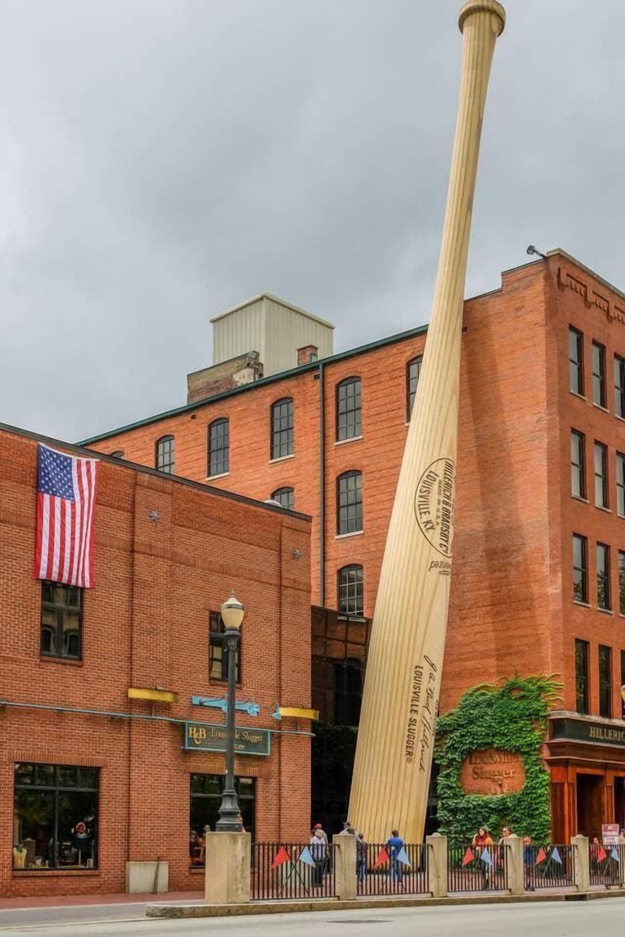 Un bate de béisbol de madera grande frente a un edificio de ladrillo con una bandera estadounidense y un letrero de 'Hillerich & Hess'.