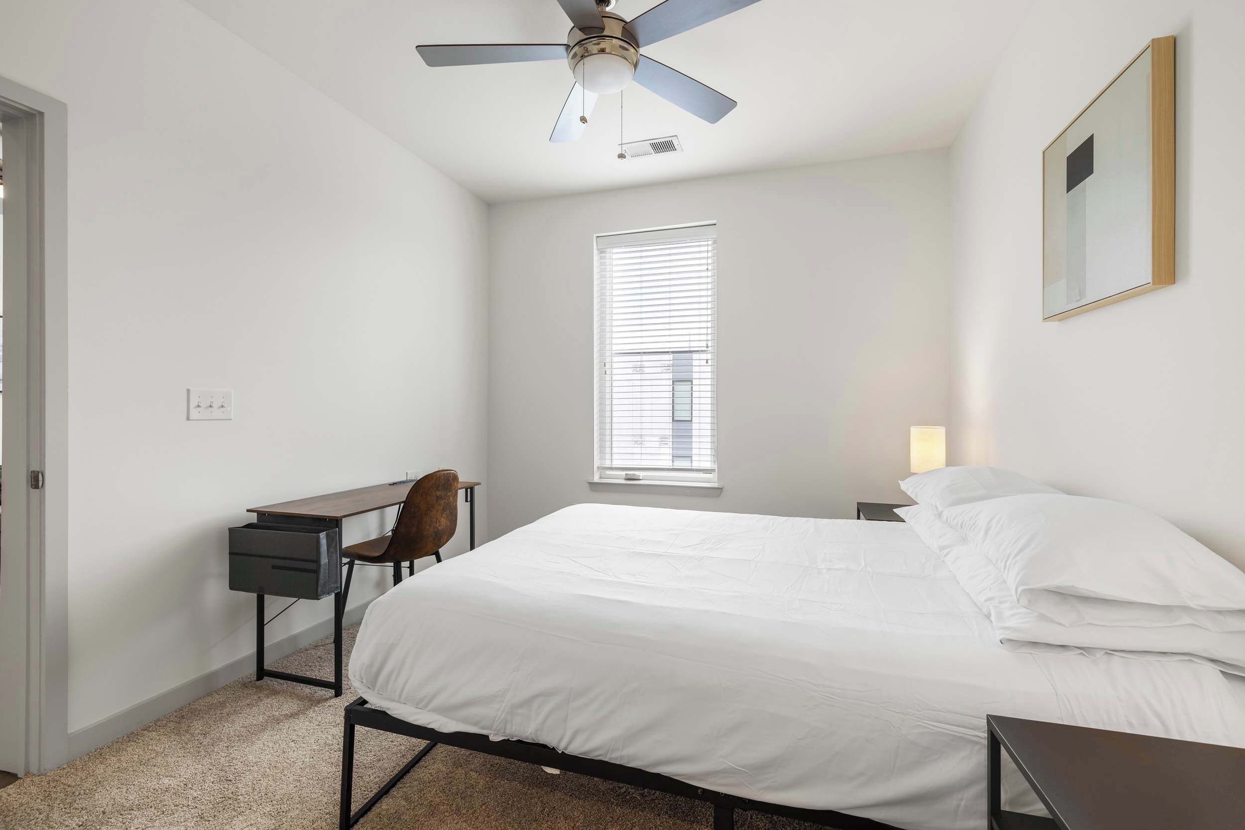 A bedroom with a bed, a desk, a chair, a ceiling fan, a window, and a wall-mounted light.