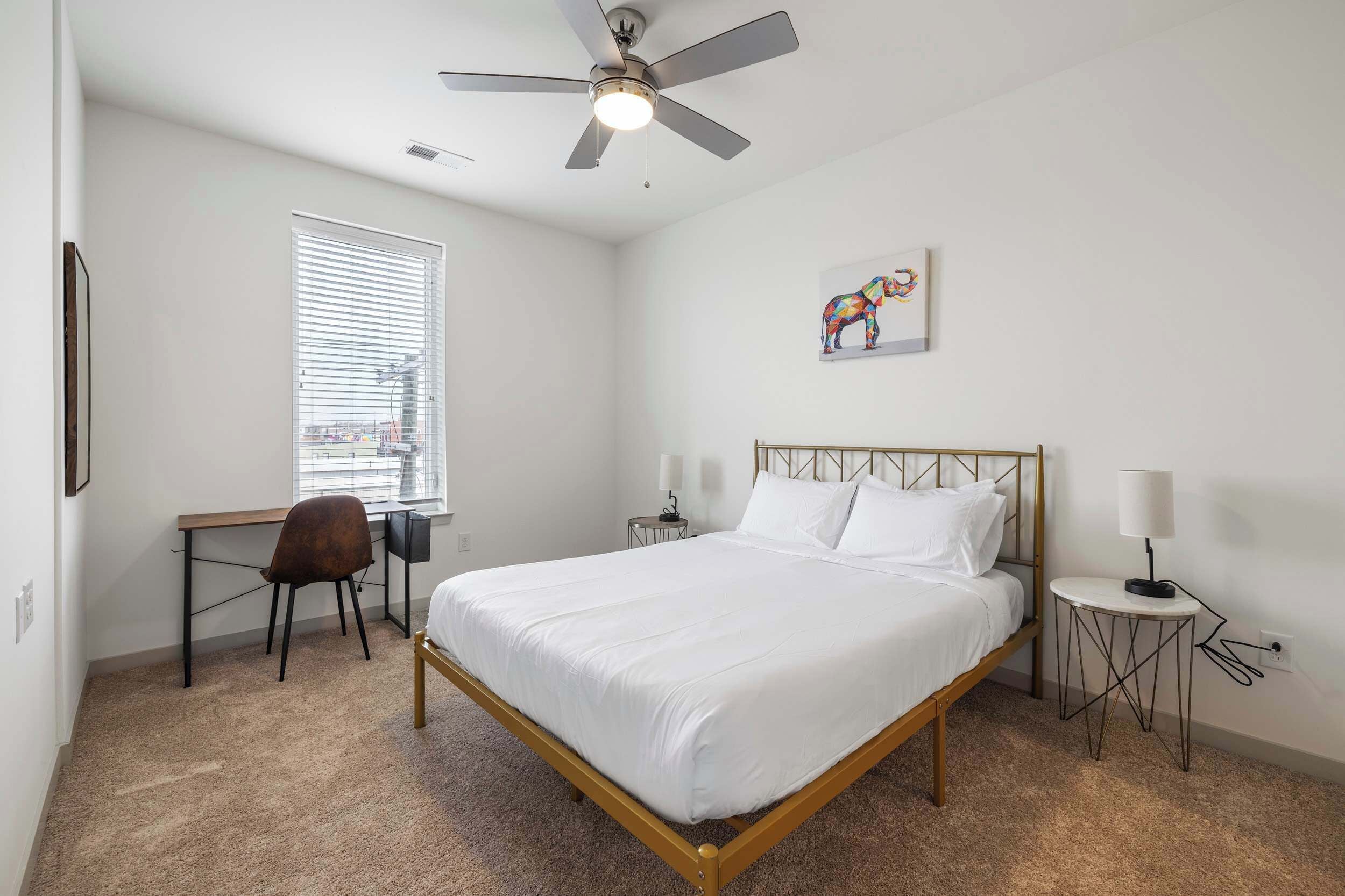 A bedroom with a bed, a desk, a chair, a ceiling fan, a window, and a wall-mounted artwork.