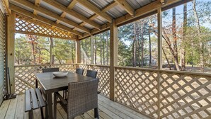 Eine überdachte Terrasse im Freien mit einem Holztisch und -stühlen, umgeben von einem Gitterzaun und Blick auf eine bewaldete Gegend.