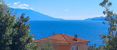 A coastal house with a red-tiled roof, a balcony, and a view of the sea and mountains.