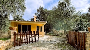 A rustic stone pathway leading to a yellow building with a tiled roof, surrounded by greenery and a wooden fence. - Cottage with large outdoor space and A/C (Aglientu)