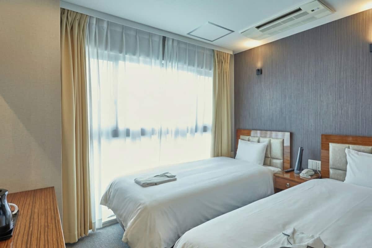 Ein Hotelzimmer mit zwei Betten, einem Holznachttisch, einer Kaffeemaschine und einem großen Fenster mit Vorhängen.