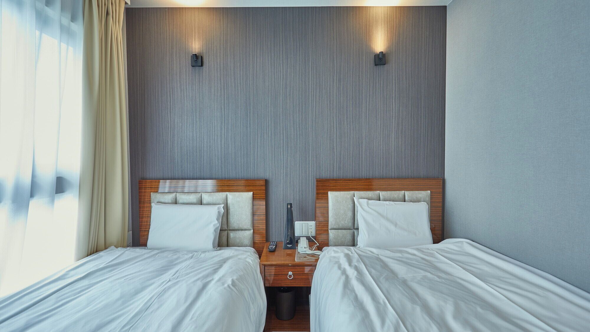 A hotel room with two beds, a wooden nightstand, a telephone, and a wall-mounted light fixture.