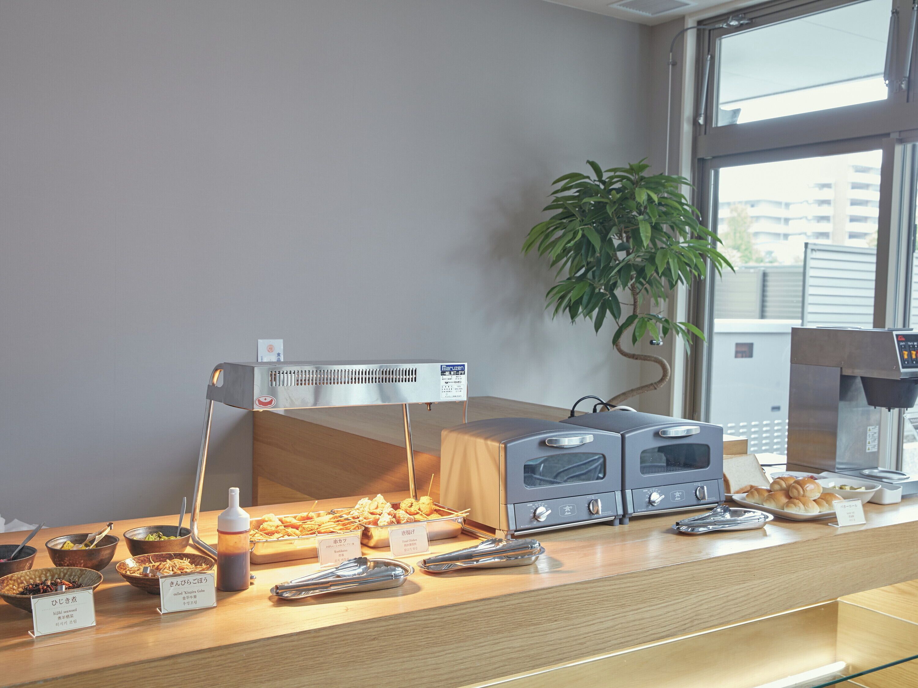 A buffet setup with various food items and utensils, a plant, and a window with a view of buildings.