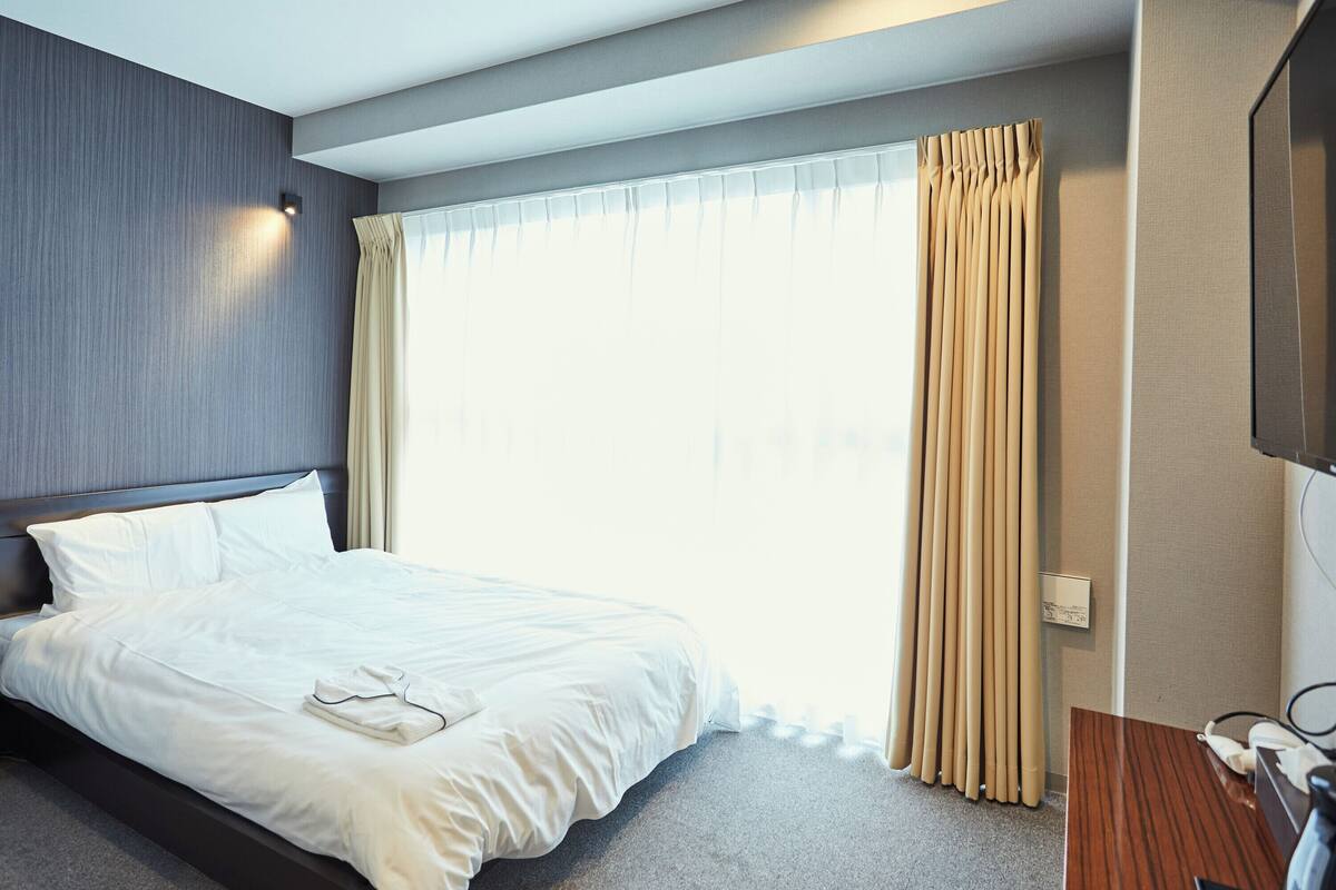 Ein modernes Hotelzimmer mit einem aufgeräumten Bett, einem großen Fenster mit Vorhängen, einem Flachbildfernseher und einer kleinen Schreibtischzeile.