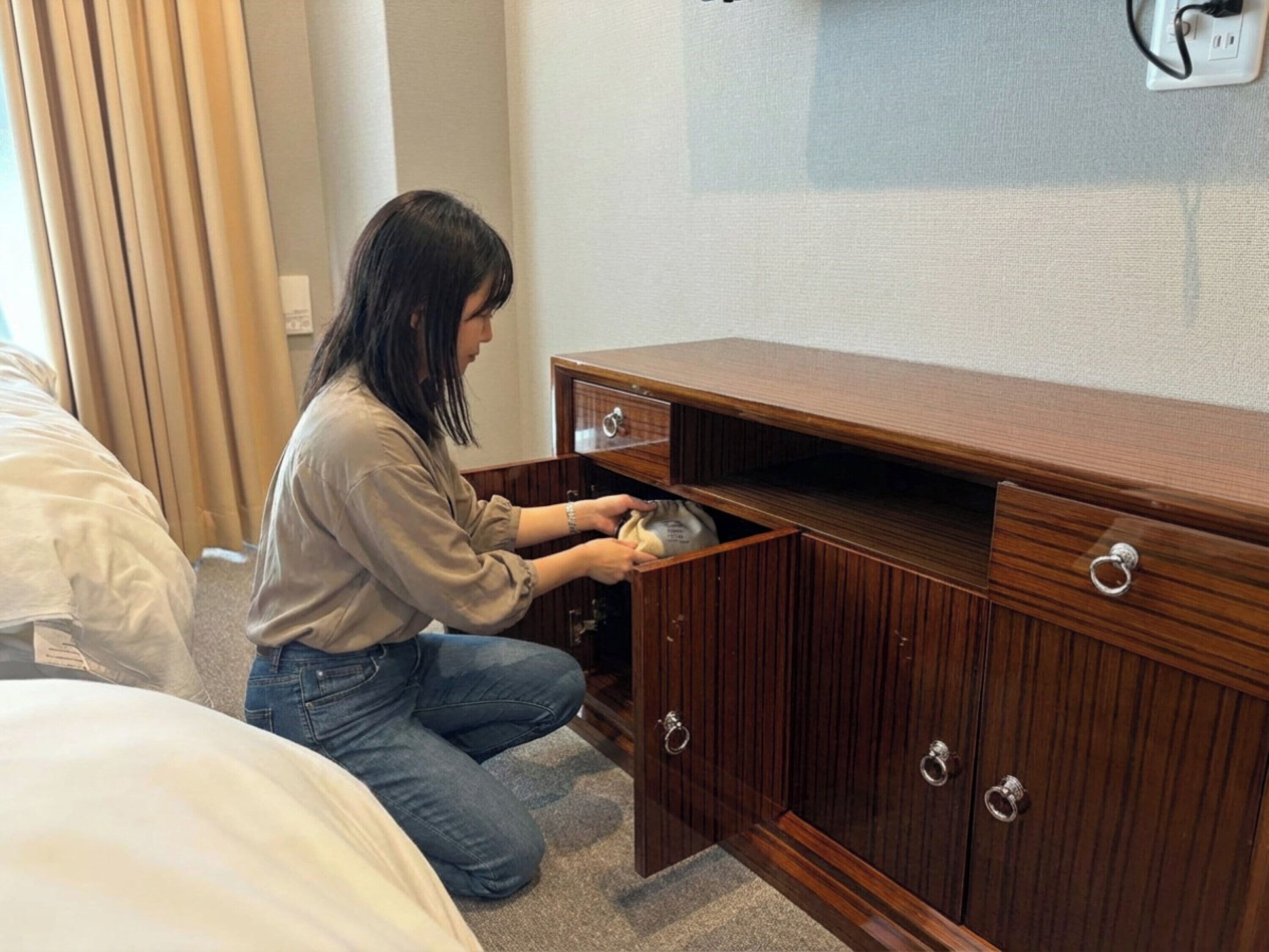 Una persona arrodillada junto a un aparador de madera con un cajón, una cama con ropa blanca y un enchufe en la pared.