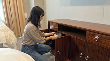 Una persona arrodillada junto a un aparador de madera con un cajón, una cama con ropa blanca y un enchufe en la pared.