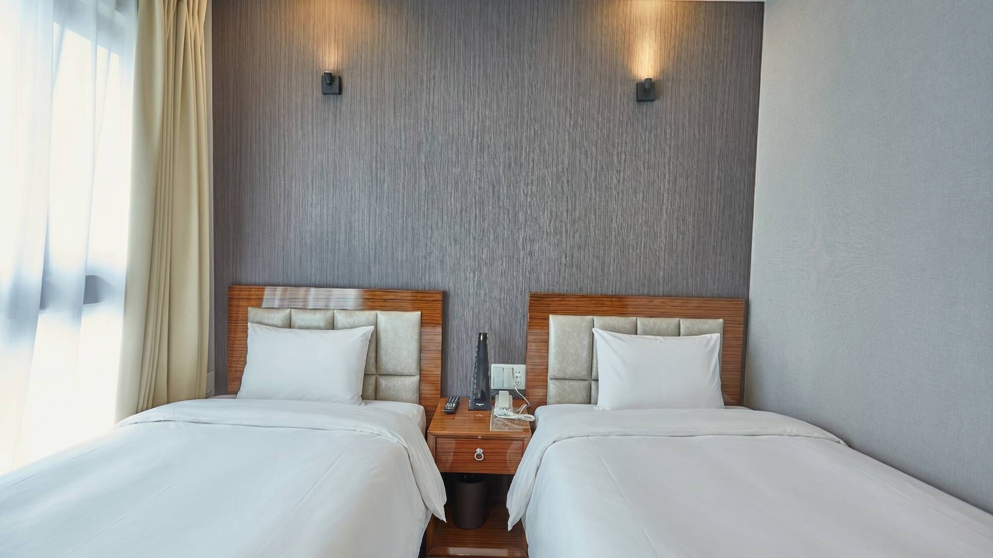 A double room with two beds, a wooden headboard, a nightstand with a phone and remote, and a wall-mounted light fixture.